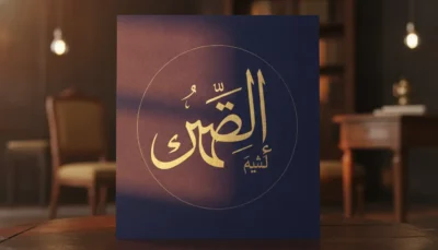 Decorative navy poster with gold Arabic calligraphy inside a circle, placed on a wooden table in a dimly lit room.